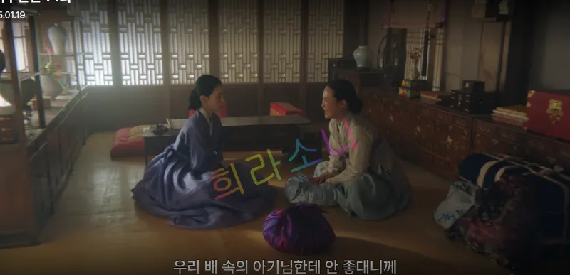 태영과 막심이 전통 가옥 내부에서 마주 앉아 대화하고 있다. 태영은 보랏빛 한복을 입고 차분하게 앉아 있으며&amp;#44; 오른쪽 막심은 하얀 저고리와 푸른 치마를 입고 있다. 주변에는 전통 목가구와 장식들이 놓여 있고&amp;#44; 창문을 통해 들어오는 자연광이 방 안을 은은히 비추고 있다. 자막: 