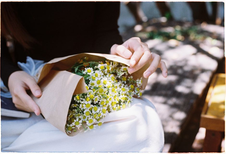 A woman tenderly holds a bouquet of chamomile flowers wrapped in brown paper while sitting outdoors.