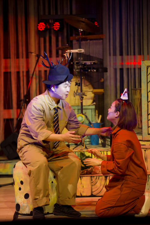 어린이극 스타일의 무대에서 뿔 머리띠를 쓴 남자 배우와 고양이 귀 머리띠를 쓴 여자 배우가 극적으로 대화하는 장면