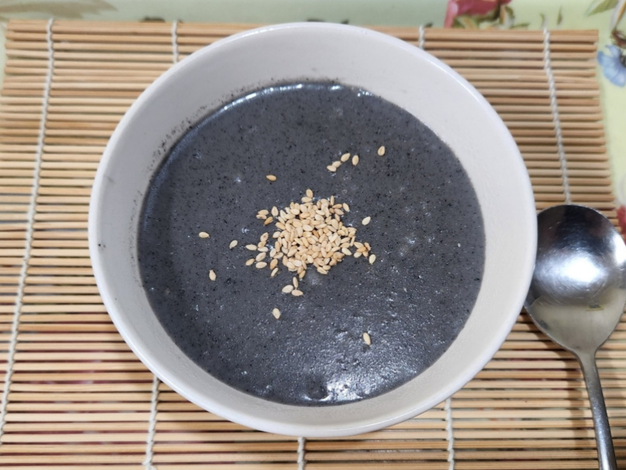 검은깨를 갈아서 밥과 함께 끓여서 만든 검은 깨죽을 동그랗고 하얀 그릇에 반쯤 담아서 대나무 깔판 위에 쇠숟가락과 함께 놓아두고 찍은 사진