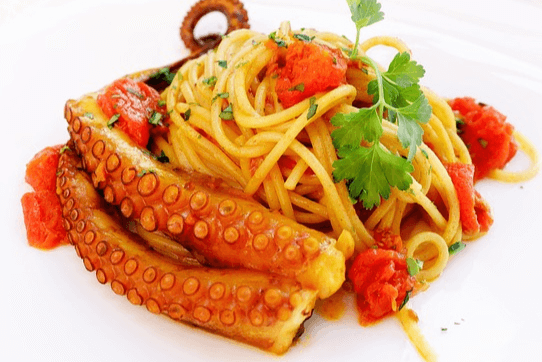 하얀 접시에 문어 다리가 있는 스파게티가 있다. 볶은 방울토마토와 초록 채소가 얹어진 모습이다.