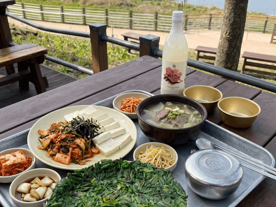대구 달성군 화원유원지 사문진주막촌 야외 나무 테이블 위에 부추전과 두부김치, 소고기국, 막걸리 한 병과 김치, 콩나물, 무채 반찬이 쟁반에 차려져 있고 뒤편에 나무와 벤치가 보이는 모습