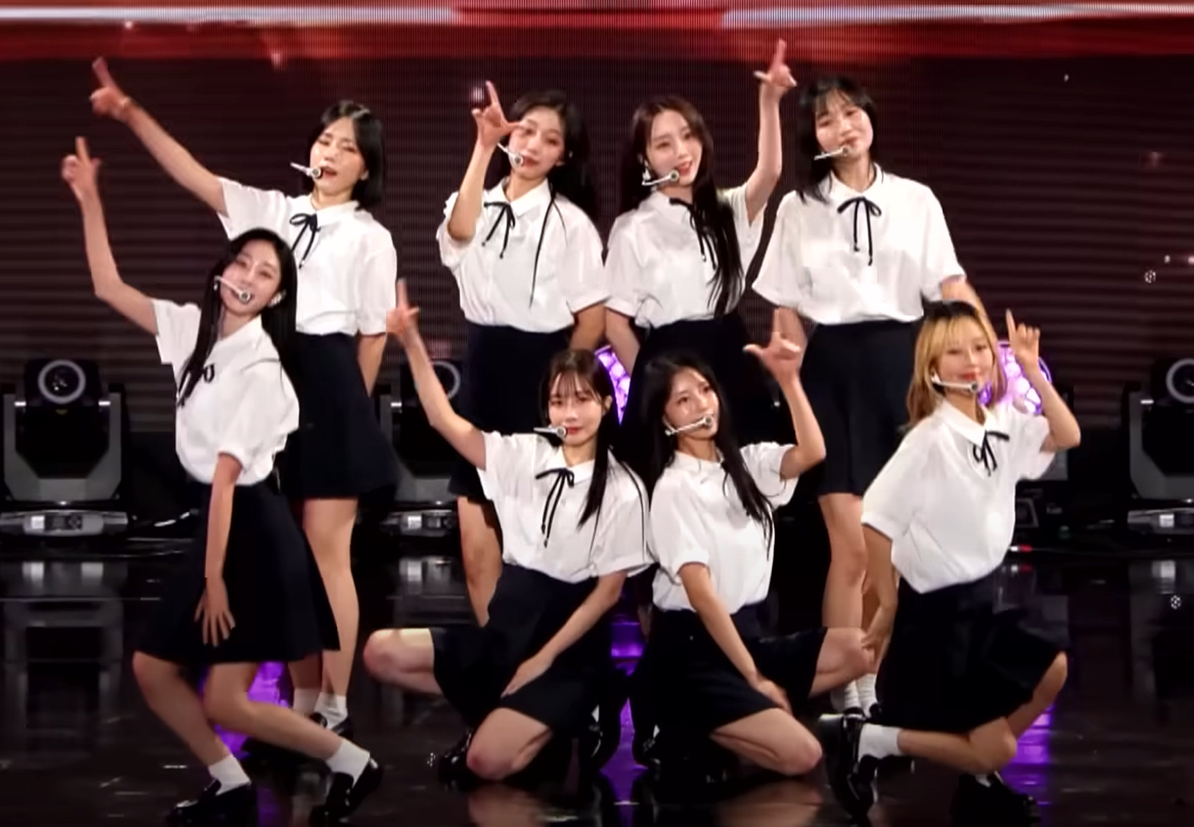 러블리즈의 8명 멤버가 무대 위에서 흰 셔츠와 검은 치마를 입고 공연을 하고 있다. 모든 멤버들이 포즈를 취하며 밝게 웃고 있다