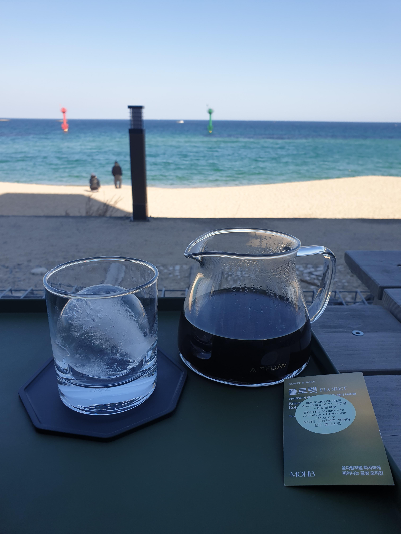 바다가 보이는 테이블 위에 커피가 담긴 서버와 얼음이 담긴 컵이 놓여있다.