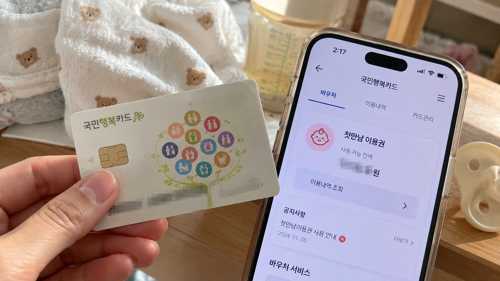 국민행복카드를 손에 들고 스마트폰에서 첫만남이용권 바우처 잔액과 이용내역 조회 화면을 확인하는 모습