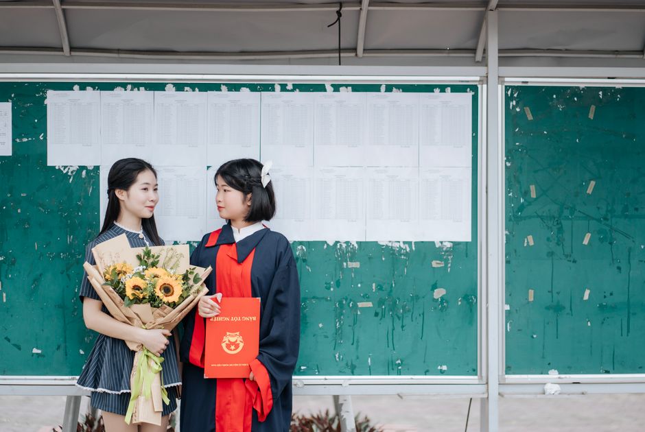 Two graduates celebrate in front of a bulletin board with a sunflower bouquet and diploma.