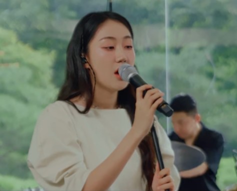 허회경이-하얀-옷을-입고-마이크를-오른손은-마이크를-잡고-왼손은-마이크대를-잡고-노래를-부르고-있는-사진