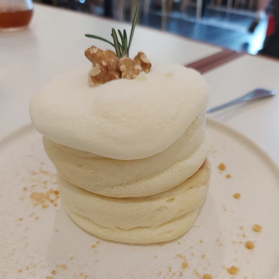 흰 접시 위에 3단으로 쌓인 부드러운 수플레 팬케이크 위에 생크림과 호두, 로즈마리가 장식된 모습