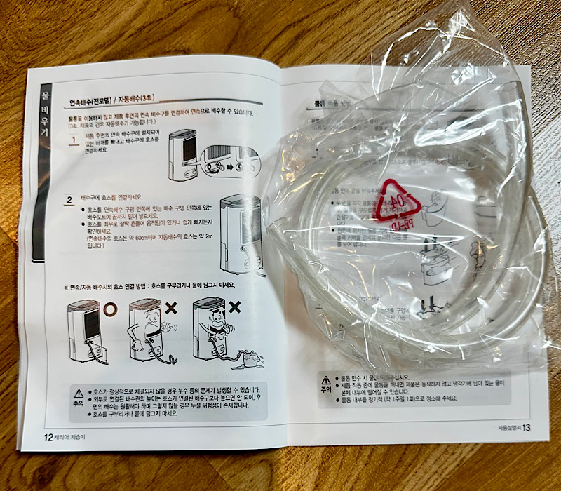 캐리어 제습기 설명서 12~13페이지 위에 연속 배수용 호스가 비닐에 포장된 상태로 놓여 있으며, 제품 연결 방법과 주의사항 삽화가 함께 인쇄되어 있음