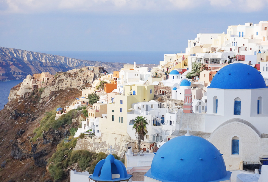 그리스 산토리니 섬으로 파란색 둥근 지붕, 하얀색 벽의 집들과 절벽 아래 바다가 보인다.