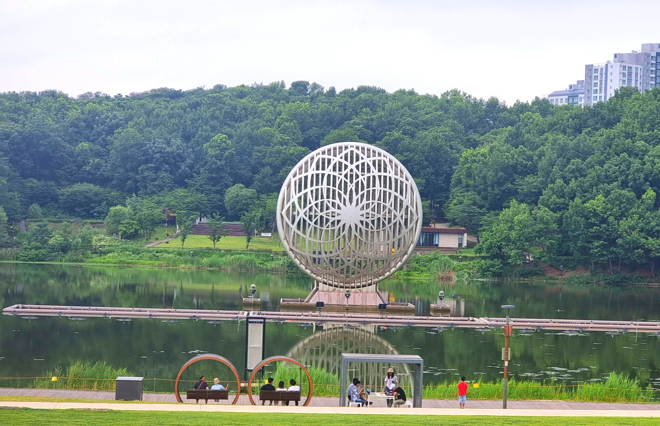 호수공원 달 모양 조형물과 테이블과 그네에 사람들