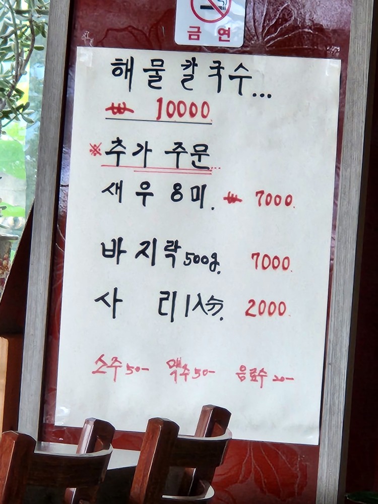 '해물칼국수 10000'이라고 정성스럽게 쓰인 손글씨 메뉴판. 새우, 바지락 등 추가 메뉴도 보인다.