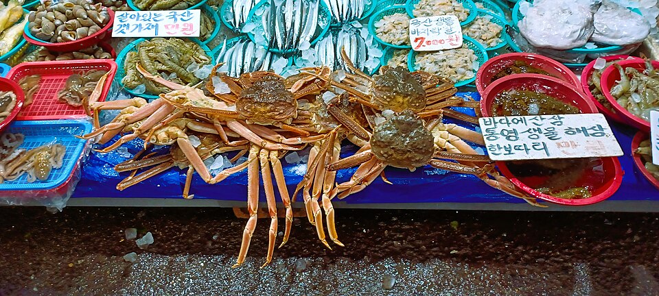 Crabs piled on a seafood counter at Mangwon Market, surrounded by colorful trays, handwritten Korean signs and fish on ice.