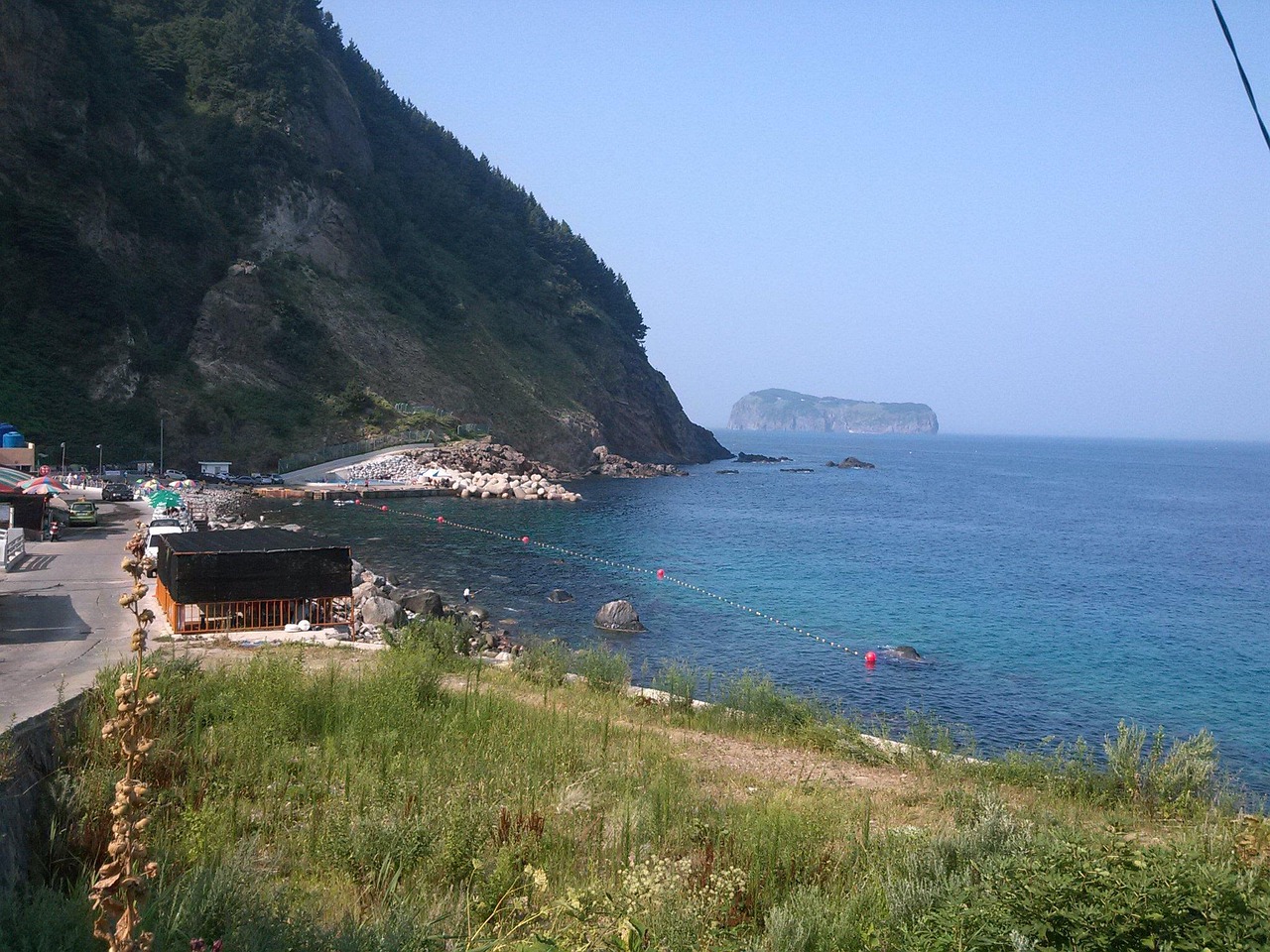 깎아지른 듯한 해안 절벽과 푸른 바다가 맞닿은 조용한 어촌 마을. 맑고 투명한 바다 너머로는 완만한 언덕 모양의 섬이 멀리 떠 있으며, 방파제와 작은 부두가 아담하게 자리잡아 있다. 해안선을 따라 줄지어 있는 천막과 건물들은 마을의 소박한 삶의 흔적을 보여준다. 해안도로 옆으로는 야생초가 무성하게 자라고, 전체적으로 자연과 인간의 삶이 조화롭게 공존하는 평화로운 풍경을 담은 사진.