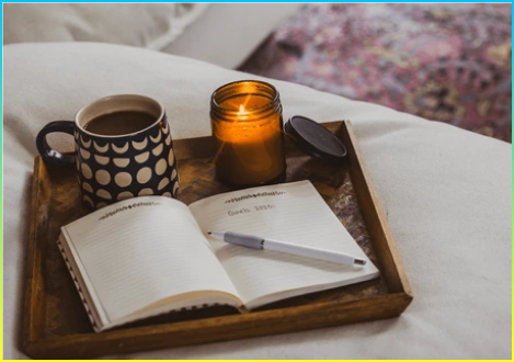 커피 한 잔, 촛불, 그리고 흰색 소파 위에 놓인 쟁반에 놓인 노트와 일기가 새해의 목표를 적는 아늑한 거실 장면.