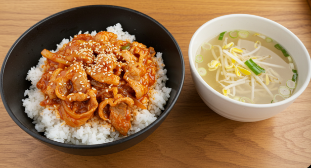 붉은 양념의 제육볶음이 밥 위에 먹음직스럽게 얹어져 있고, 그 위로 통깨가 뿌려져 있다.