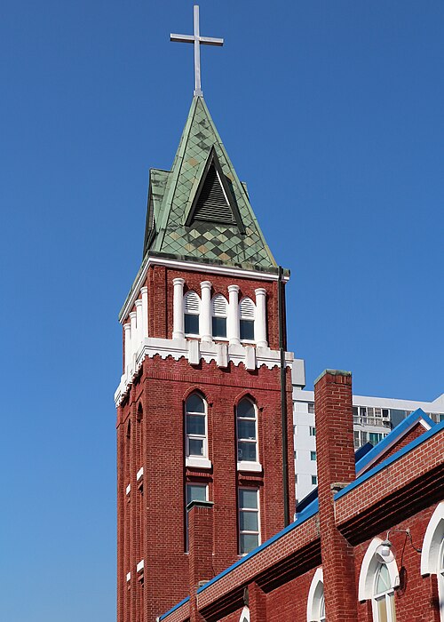 Yangnim Church&rsquo;s red-brick tower rises to a green, cross-topped spire, with white arched details and blue roof edging against a clear sky.