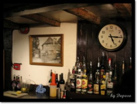 영국 런던 근교의 한 펍 내부 바 벽면. 나무 들보가 드러난 천장 아래, 다양한 술병들이 진열되어 있으며, 벽에는 둥근 시계와 고풍스러운 흑백 건물 사진 액자가 걸려 있다. 아늑하고 전통적인 영국 펍의 정취가 느껴진다.