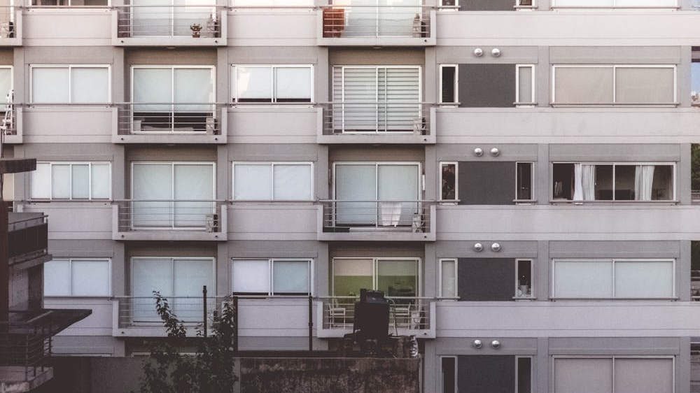 옅은 회색 콘크리트로 지어진 아파트 건물의 정면. 여러 층에 걸쳐 비슷한 크기의 흰색 창문과 금속 난간이 설치된 작은 발코니들이 반복적으로 배열되어 있으며, 밀도 높은 공동 주택의 모습을 보여줍니다.