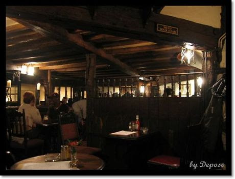 영국 런던 근교의 전통적인 펍 내부. 낮은 천장에는 나무 들보가 드러나 있고, 손님들이 테이블에 앉아 식사와 대화를 즐기고 있다. 앞쪽 테이블 위에는 케첩과 머스타드 병, 냅킨이 놓여 있으며, 따뜻하고 아늑한 조명이 공간을 부드럽게 감싼다.