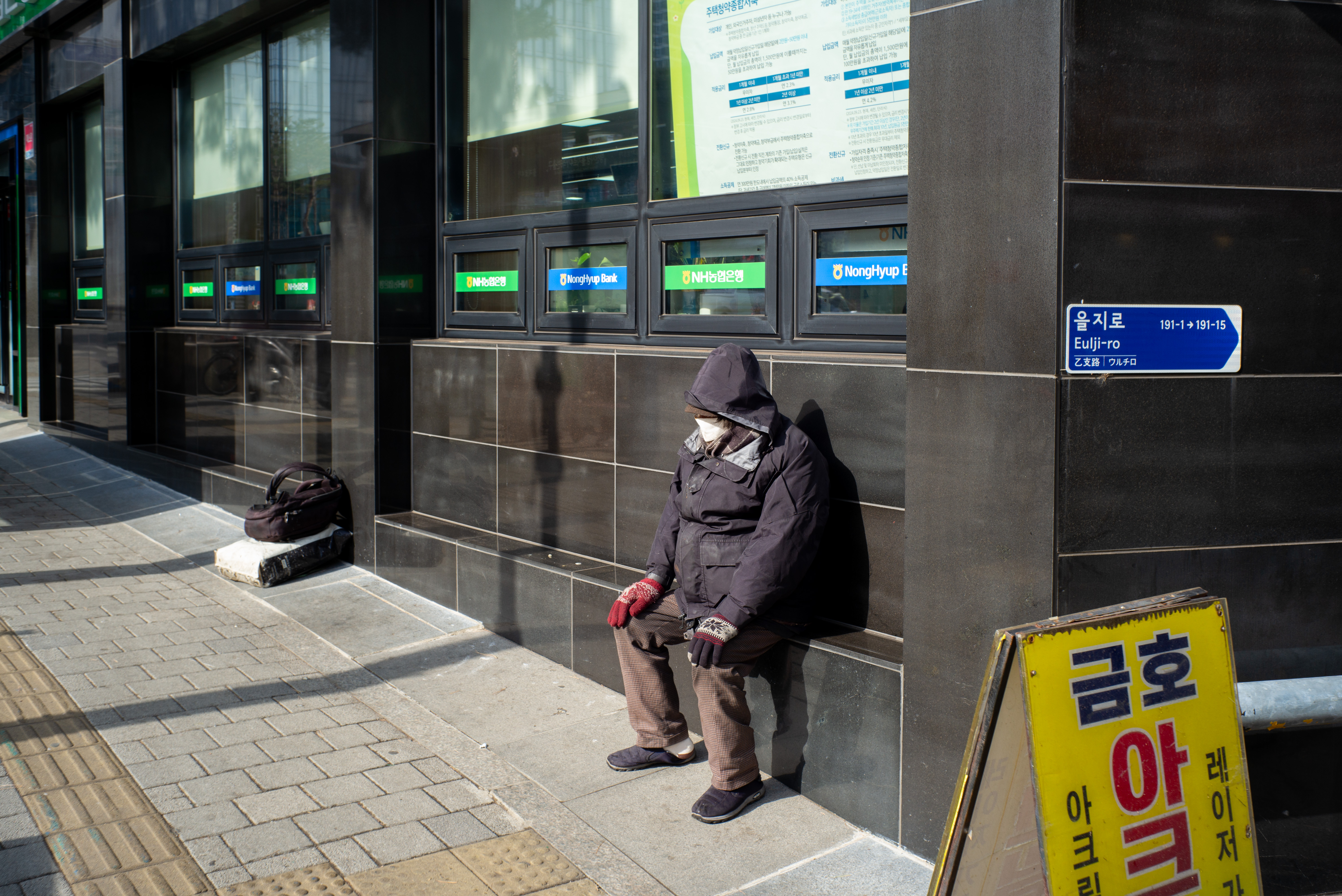을지로 길거리 검은색 대리석 건물 외벽 아래 의자에 앉아 휴식을 취하고 있는 마스크를 쓴 노인