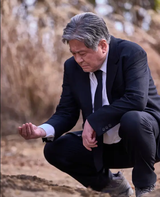 파묘- 흰와이셔츠 검은넥타이 검은 양복을입고 쪼그리고 앉아 오른손에 흙을 들고 보고있는 최민식