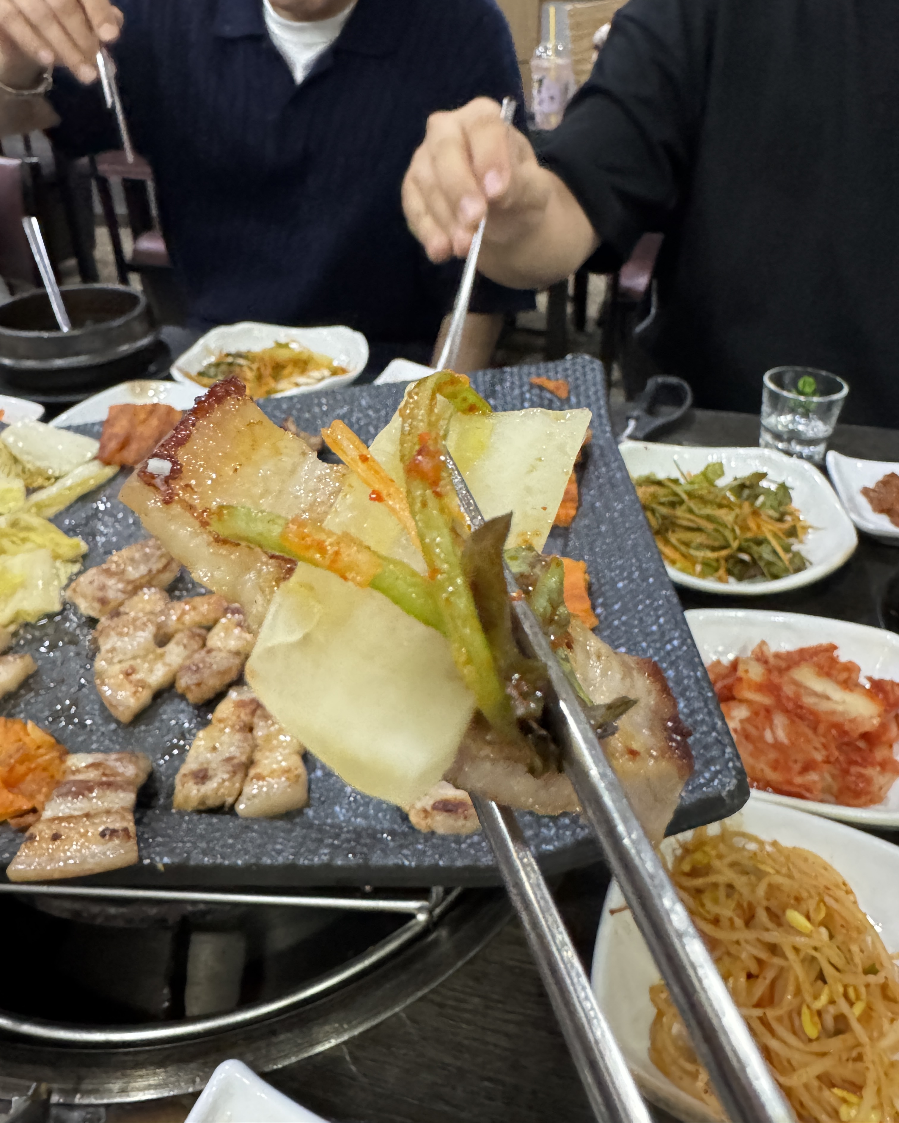 구운 오겹살에 백김치와 파채무침을 올려 한입 크기로 만든 조합을 젓가락으로 든 모습.