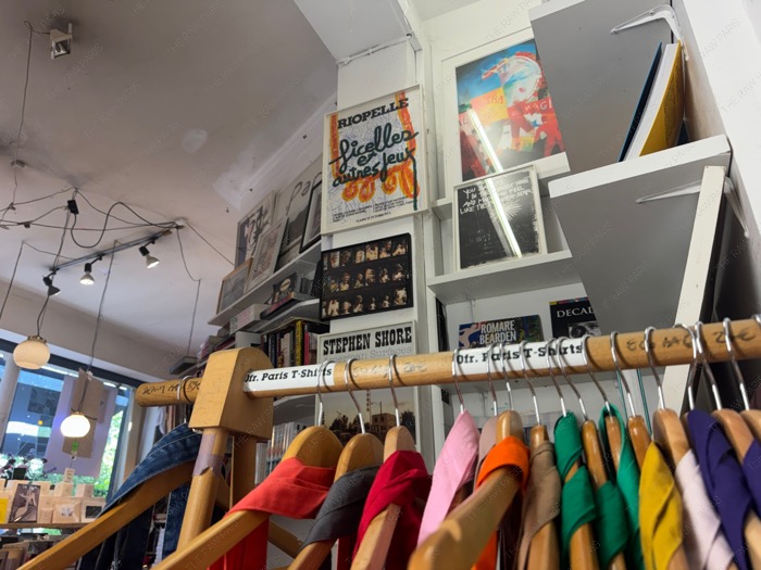Colorful graphic t-shirts hanging on a wooden rack labeled Ofr. Paris T-Shirts with art posters in the background ❘ Ofr. Paris