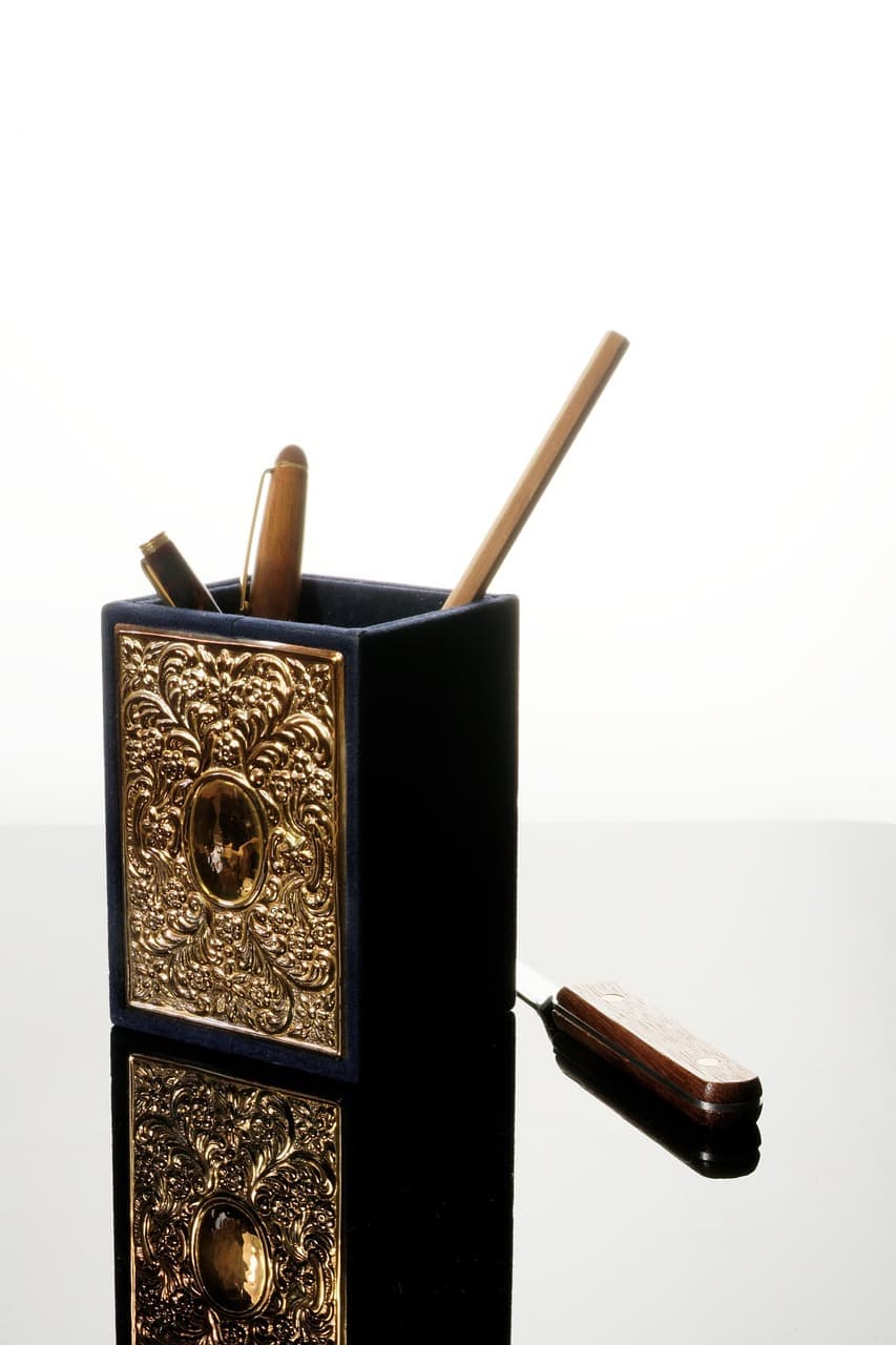 정교한 금색 장식이 새겨진 펜꽂이에 고급스러운 만년필과 펜이 꽂혀 있다. 전반적으로 클래식하고 우아한 느낌을 주는 문구류 아이템이다.
