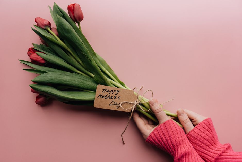 A hand holds a bouquet of tulips with a Happy Mother's Day tag on a pink background.