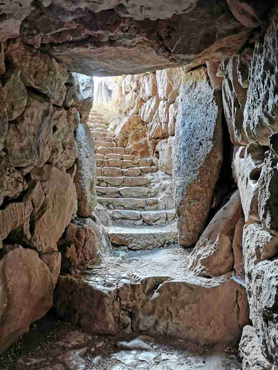 거친 돌로 쌓아올린 벽과 천장 사이로 이어지는 좁은 돌계단이 바깥쪽 밝은 빛으로 향해 나 있는 미케네 유적지의 고대 통로