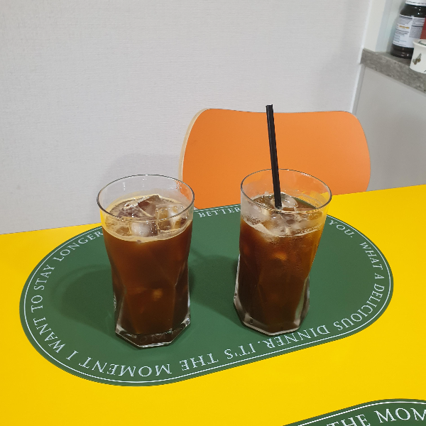 노란 테이블 위 헌터색 매트가 깔려 있고 그 위에 아이스 아메리카노가 담긴 유리잔 두 개가 있다.