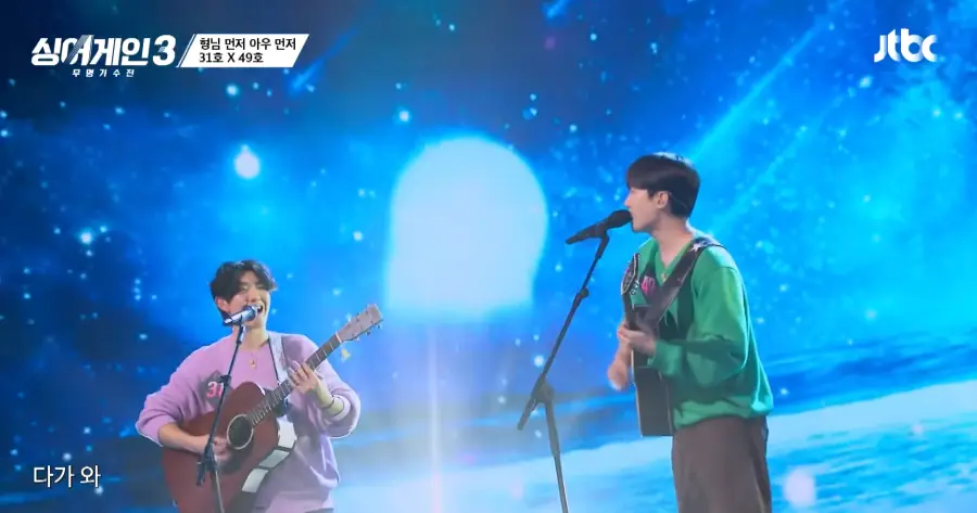 싱어게인3 - 좌측 보라색 티를입고 스탠딩 마이크 앞 갈색 기타를 치며 노래하는 31호와 우측 초록색티에 검은바지를 입고 기타를 치며 스탠딩 마이크앞에서 노래하는 49호