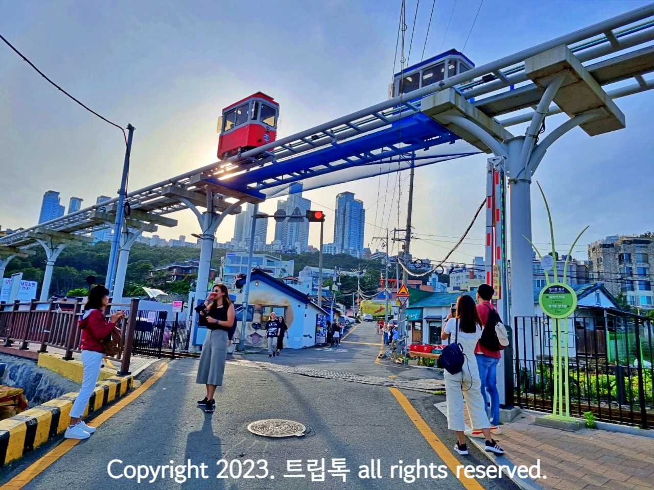 사거리 위에 높은 레일이 있고 그위를 스카이 캡슐이 지나가고 길에서 사람들이 구경도하고 사진도 찍는다.