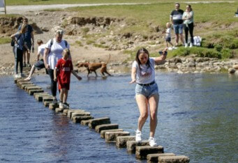 큰-폭의-강에-놓여있는-징검다리를-사람들이-줄을지어-건너는데-맨앞-여성이-두-손을-들며-균형을-맞추며-불안하게-건너고있는-모습