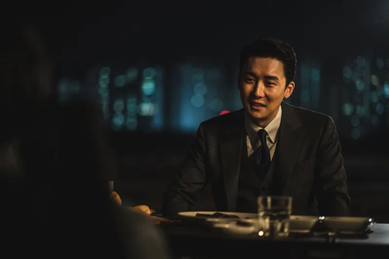 어느 술집을 배경으로 양복을 입고 소주잔이 있는 테이블에 앉아 있는 영화 보호자의 성준