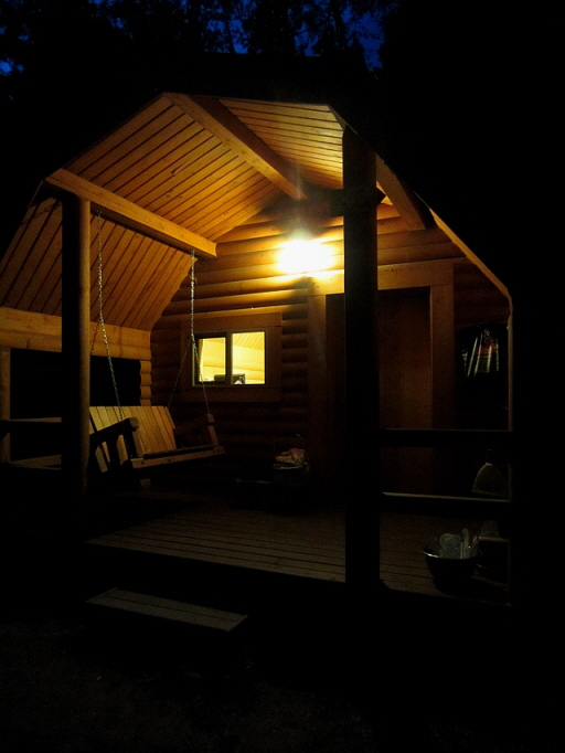 밤의 숲 속 통나무 캐빈, 불빛 아래 흔들의자와 테라스가 따뜻하게 빛남.