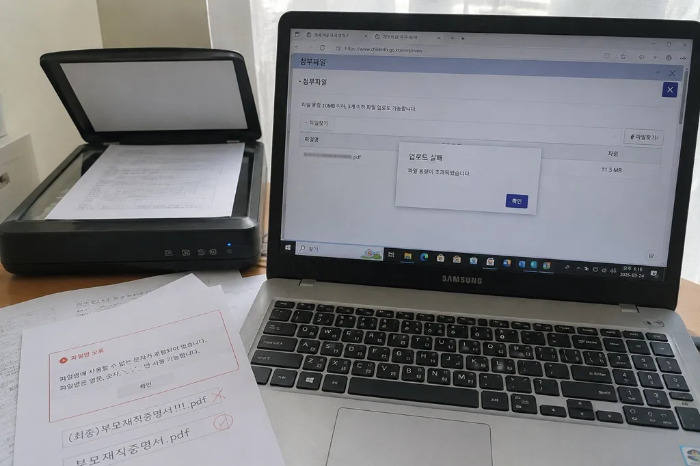 삼성 노트북 화면에 어린이집 관련 웹사이트의 첨부파일 업로드 실패 팝업과 파일 용량이 초과되었다는 안내가 떠 있는 모습, 노트북 왼쪽에는 스캐너가 있고 그 앞에는 파일명 오류 예시와 특수문자가 제외된 올바른 파일명 예시가 적힌 인쇄물이 놓여 있는 책상 풍경