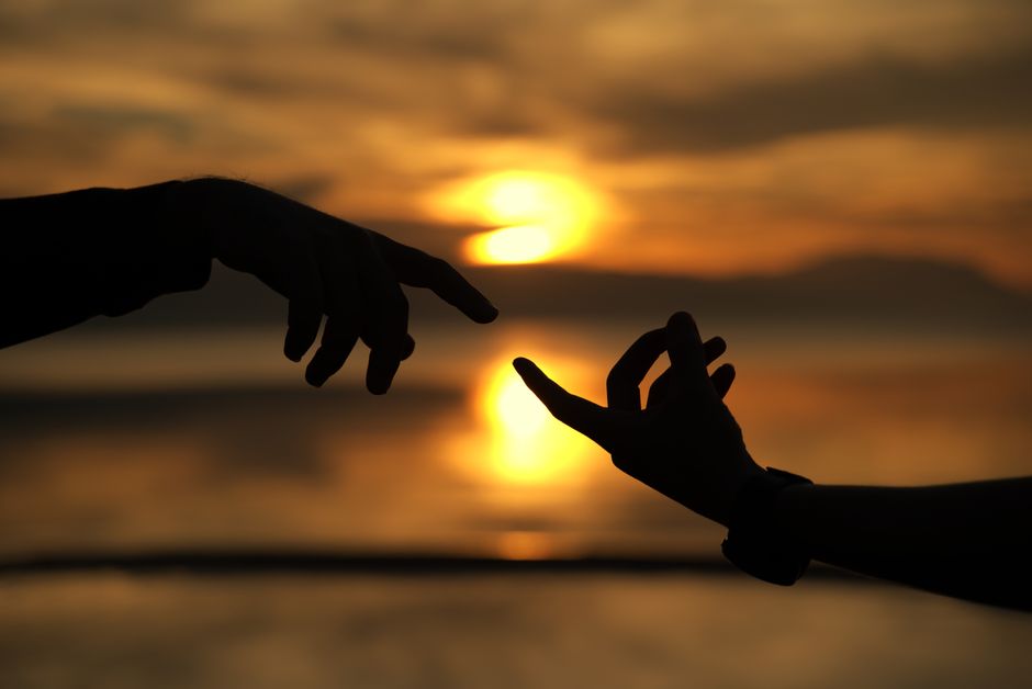 Silhouetted hands reaching towards each other against a stunning sunset background over the ocean.