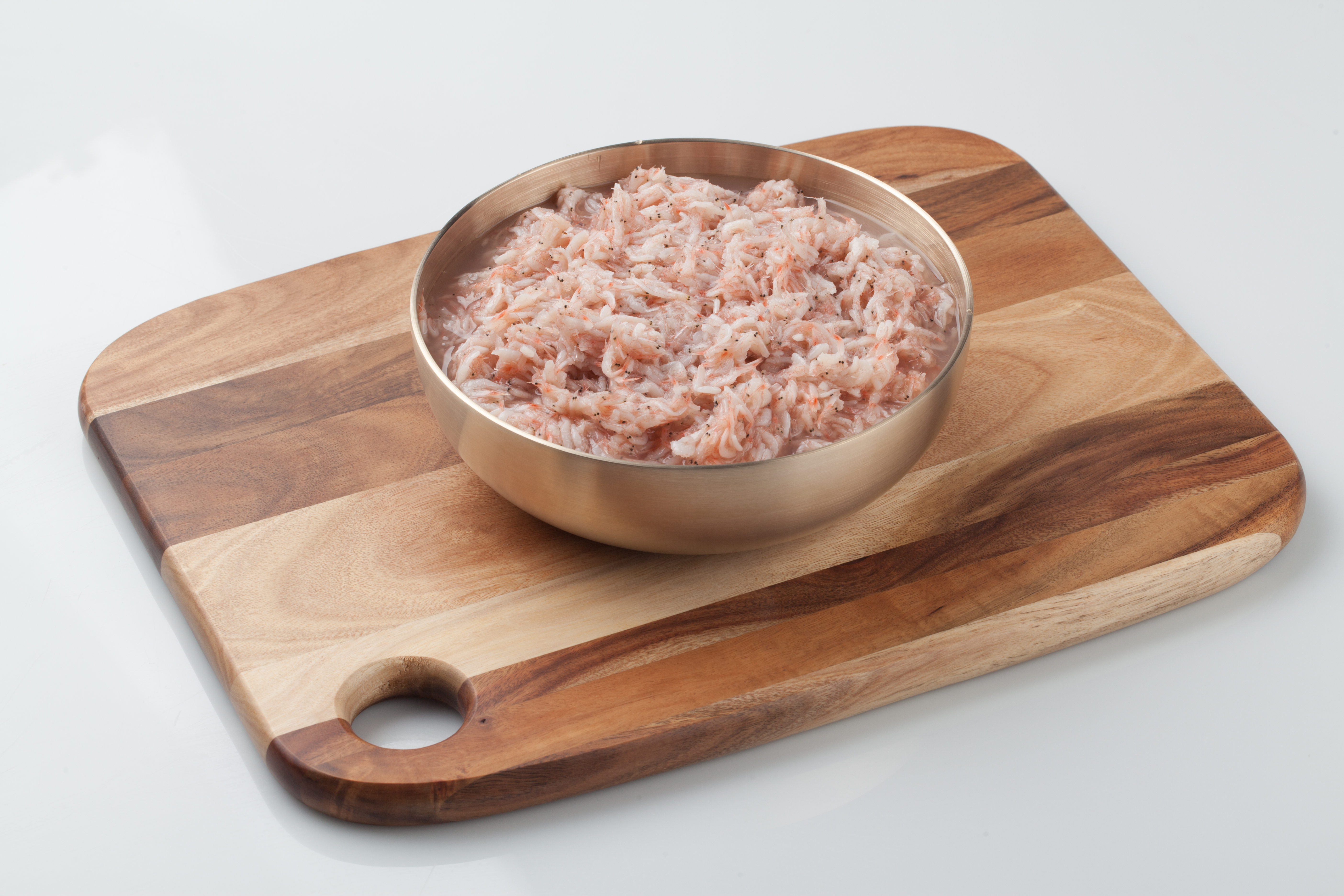 하얀 바닥 위에 네모나고 한쪽 모서리 부분에 동그란 구멍이 난 나무 도마를 두고 그 위에 쇠로 된 동그란 그릇을 두고 그 속에 새우젓을 가득 담아두고 찍은 사진