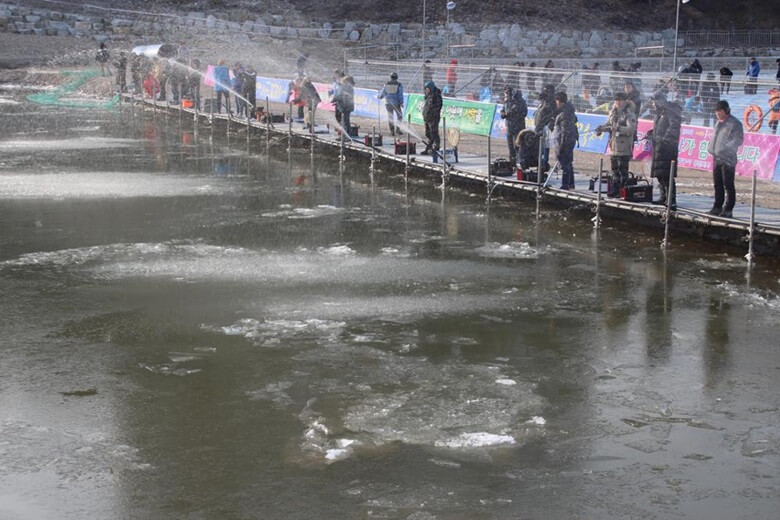 덜 얼어붙은 강을 앞에 두고 수많은 사람들이 강 주변에서 얼음 낚시를 하기 위해서 낚시대와 낚시 도구 가방을 챙겨서 서있는 모습