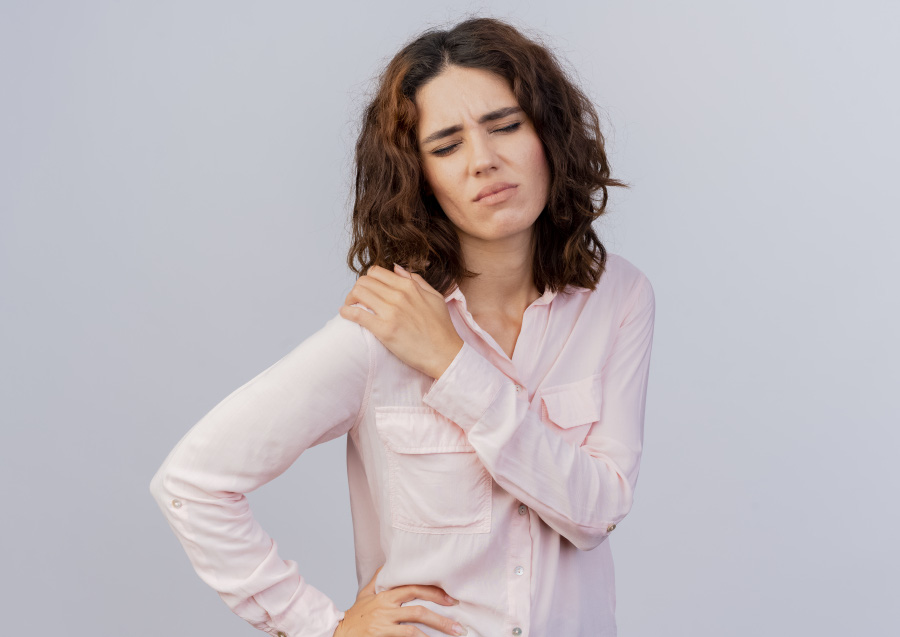 분홍색 윗옷을 입은 서양의 젊은 여성이 오른쪽 어깨 통증을 호소하며 왼쪽 손으로 통증 부위를 마사지 하며 괴로워하고 있는 것을 찍은 사진