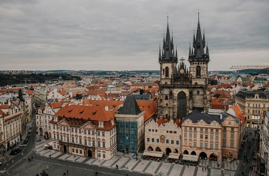 높은 곳에서 내려다본 체코 프라하 구시가지 광장의 모습. 화면 오른쪽에 두 개의 검은 고딕 양식 첨탑이 돋보이는 틴 성모 교회가 솟아 있고, 광장을 둘러싼 고풍스러운 건물들과 주황색 붉은 지붕들이 펼쳐진 풍경.