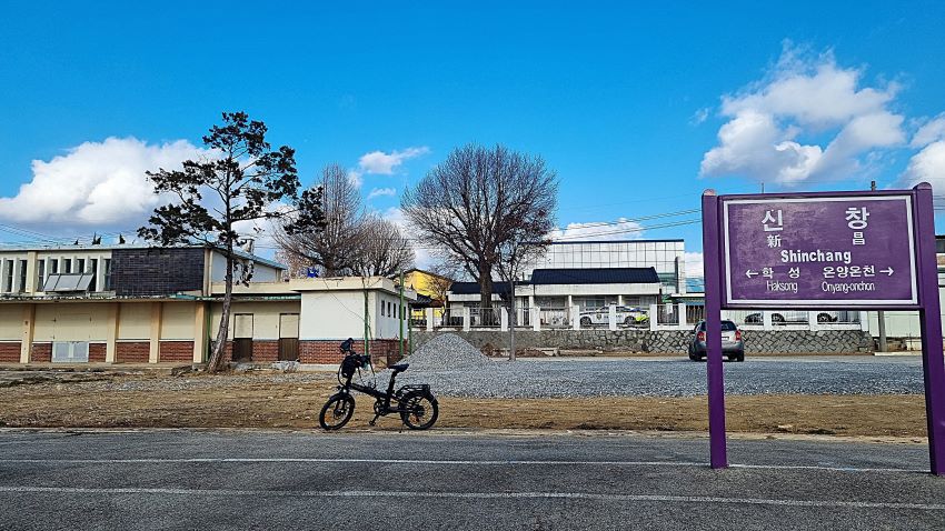 푸른 하늘, 앞에 구 신창역 역사건물, 그 앞에 나무 몇 그루, 그 앞에 세워 놓은 자전거, 우측에 보라색으로 된 역명 안내판에 한글과 영문으로 표시된 신창, shinchang, 안내판에 하행선 다음 역은 학성, 상행선 다음 역은 온양온천,