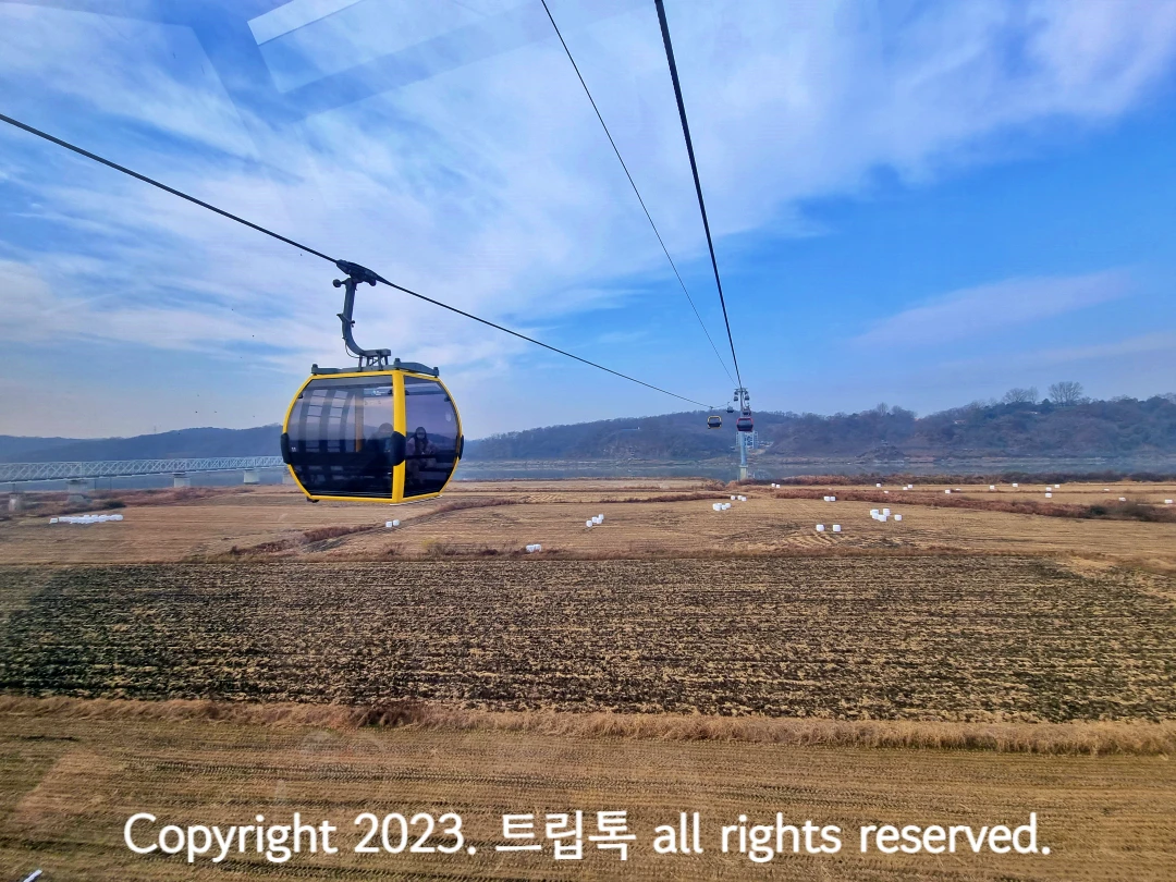 푸른하늘 아래 넓은 밭이 보이고 그 위에 노란색 둥그런 케이블카가 줄에 걸려 있다