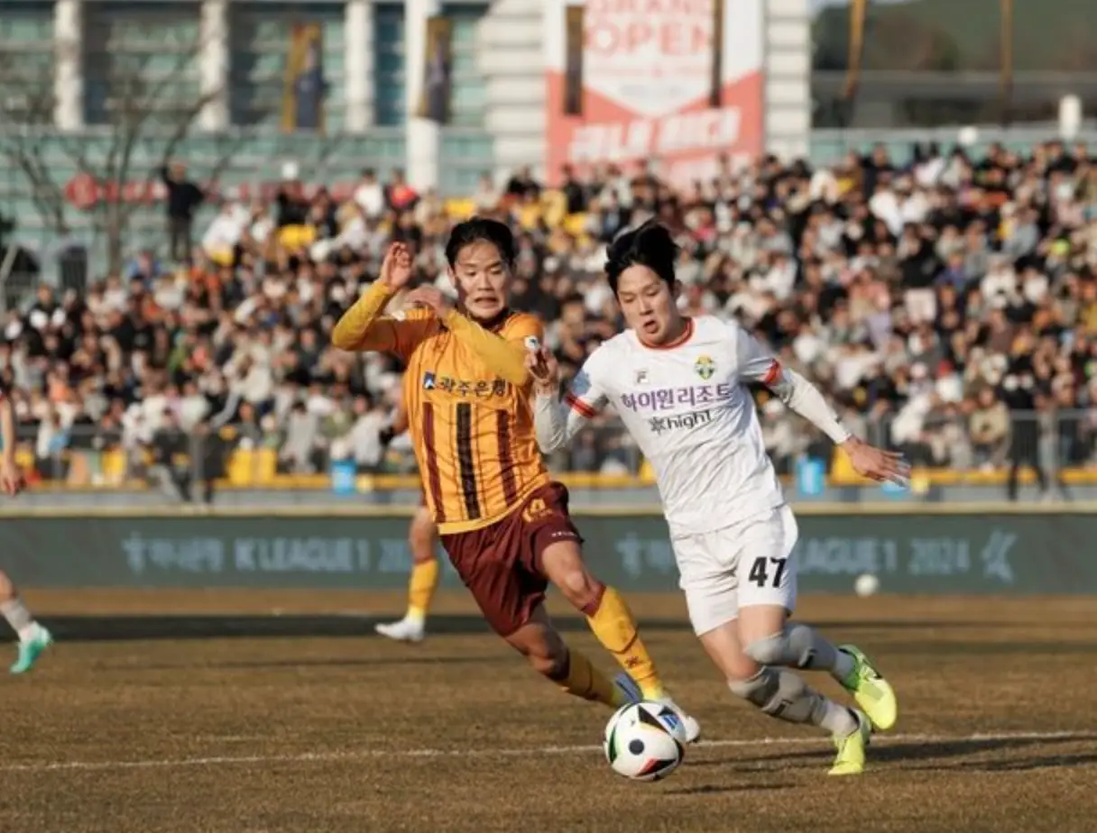 축구 경기장 흰유니폼을 입은 양민혁이 노란색과 붉은색이 섞인 유니폼을 입은 선수와 공을 서로 가로채는 모습