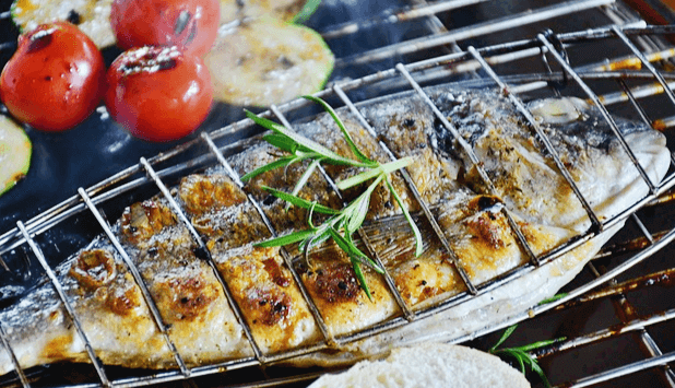 그릴에서 구워지는 도미구이 한 마리와 방울토마토 3개 및 애호박들이 있다. 도미구이 위에는 로즈마리 잎 2개가 올려져 있다.