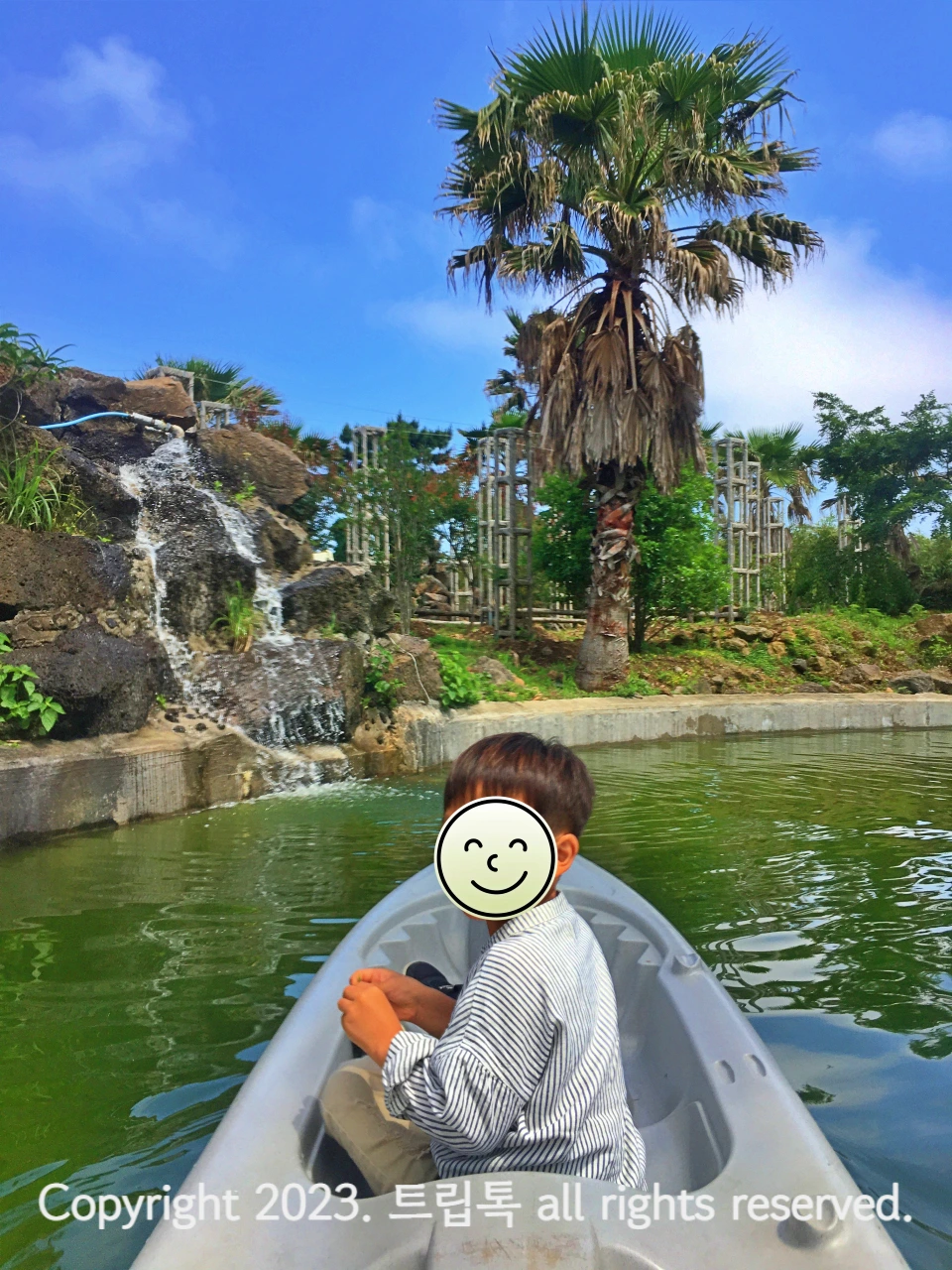 파란하늘과 폭포 조형물에 물이 떨어지고 옆에 야자가 있으며 하얀 카약을 탄 아이가 뒤를 돌아본다
