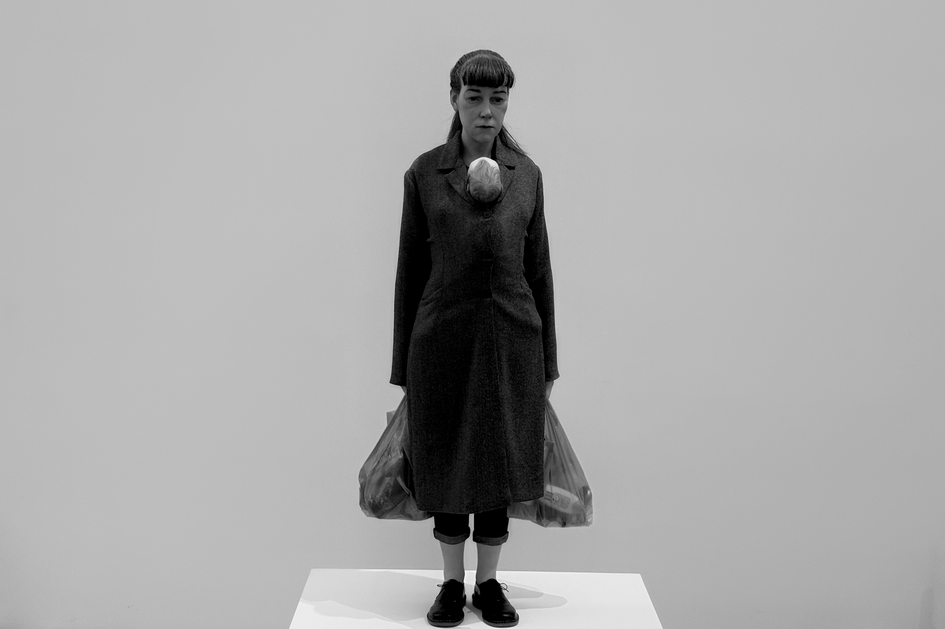 《쇼핑하는 여인》 작품 - 검은 코트를 입고 양손에 쇼핑백을 든 여성의 조각상. 무표정한 얼굴과 경직된 자세가 현대 여성의 고단함을 표현합니다. 흰색 전시 받침대 위에 서 있으며 극사실적 표현이 돋보입니다.