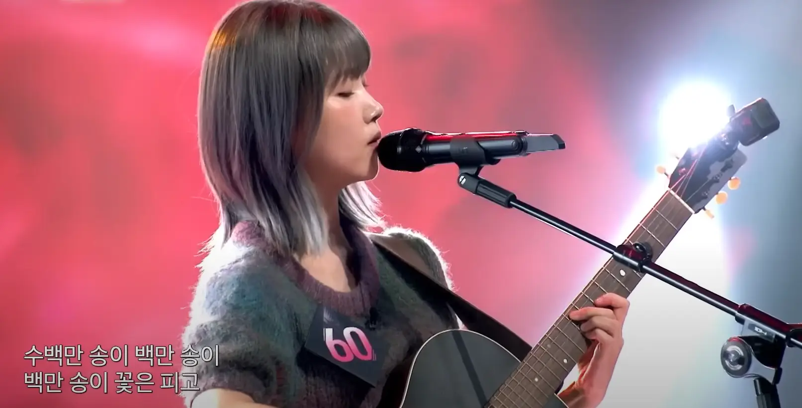 싱어게인 3- 분홍색을 배경으로 키타를 치며 가슴에 60번호표를 달고 단발머리를 늘어뜨린채 노래부르는 60호